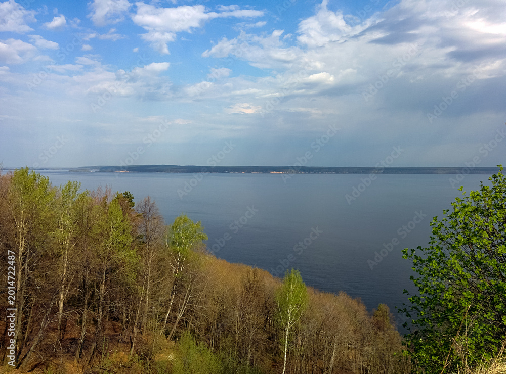 Obraz premium A scenic view of a calm lake bordered by spring trees, under a partly cloudy sky with soft sunlight