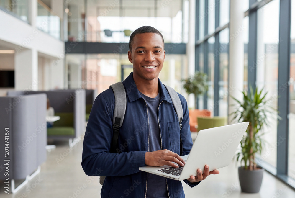 Fototapeta premium Portrait of a happy male university student holding a laptop on a modern college campus