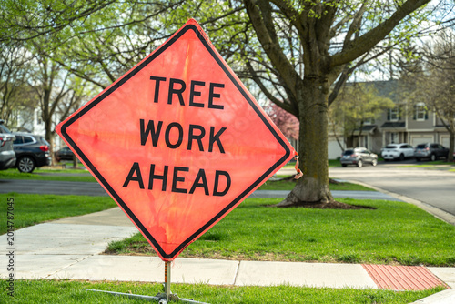 Tree work ahead sign in residential neighborhood. Warning sign about tree maintenance on suburban street with houses and cars.