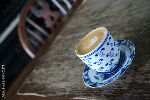A cup of latte sits elegantly on a saucer, positioned atop a weathered wooden table. The cup is decorated with a detailed blue pattern and the surface is textured with foam.
