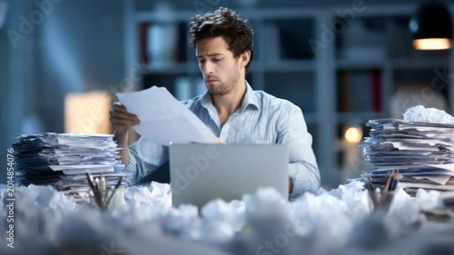 Stressed employee sitting at a messy desk with stacks of documents and throwing crumpled paper