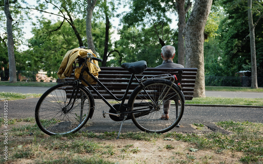 Fototapeta premium Elderly man resting on park bench beside vintage bicycle in urban park