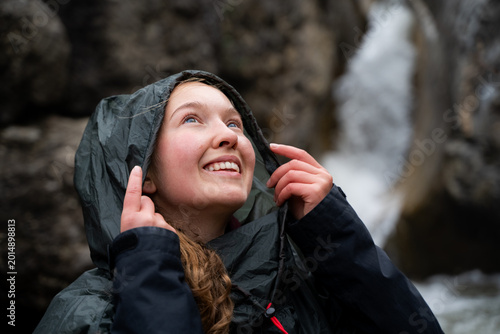 A young smiling woman is putting on a raincoat before it starts to rain outdoors, using protective clothing to protect herself from bad weather.
