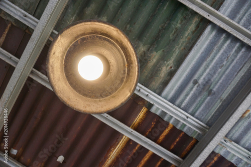 Tableau sur toile A brightly lit light bulb in an old, rusty metal lampshade, mounted on a worn corrugated iron roof, creates a very strong industrial, vintage, or rustic feel