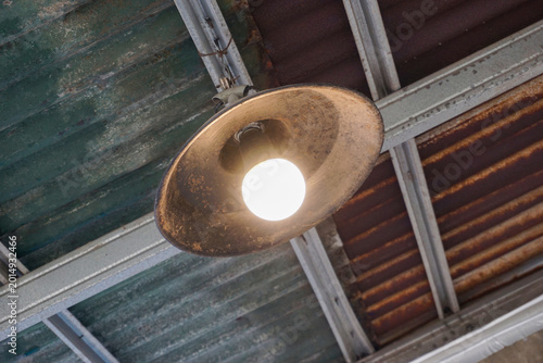 Fotografía A brightly lit light bulb in an old, rusty metal lampshade, mounted on a worn corrugated iron roof, creates a very strong industrial, vintage, or rustic feel