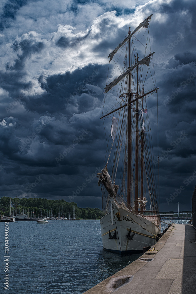 Fototapeta premium A sailing ship in the harbor of Travemünde, set against an approaching, dramatic storm front