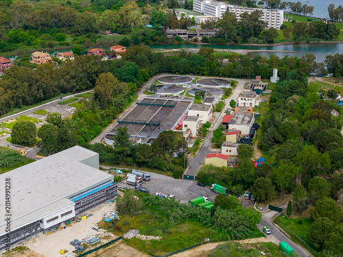 Aerial view of a wastewater treatment plant facility with clarification tanks, nearby children's traffic park, coastal landscape, and resort buildings in Corfu island, Greece