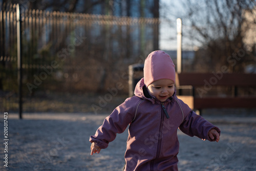 Photos smiling toddler in a pink beanie and mauve snowsuit walking in a sunlit urban pl