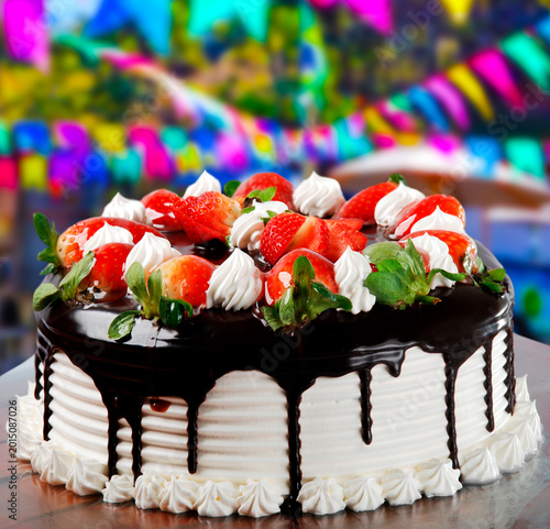 Fotografie Delicious chocolate and strawberry cake with white frosting and ganache drip, set against a vibrant, blurred Festa Junina background with colorful flags