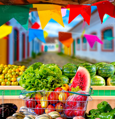 Vibrant outdoor market stall decorated with festive June party flags, featuring a shopping basket overflowing with fresh fruits and green vegetables.