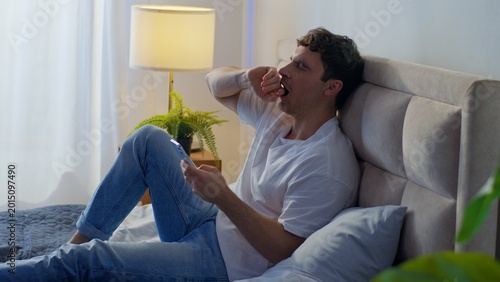 Fotografie Sleepy tired Caucasian man lying bed bedroom home yawning using mobile phone scr