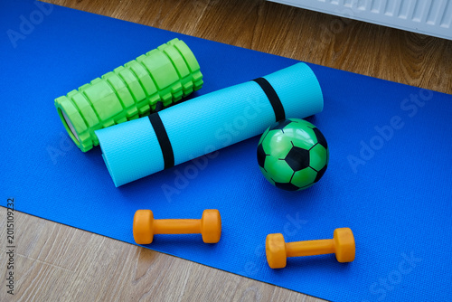 Fitness items are arranged on a blue mat in a home workout space with weights, a foam roller, and a soccer ball