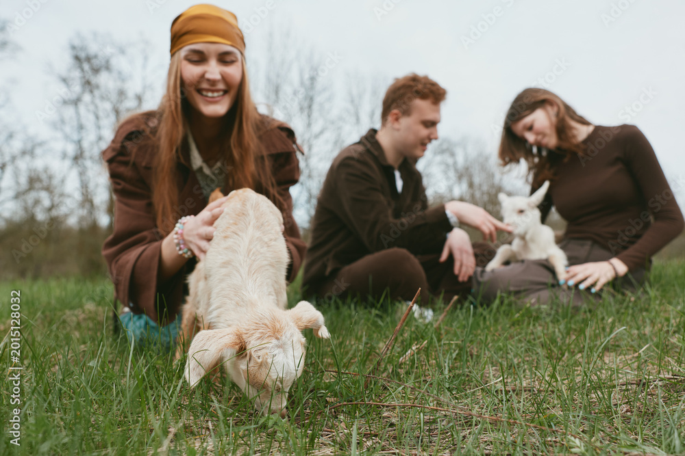 Fototapeta premium Three friends enjoy a sunny day in the field with goat, goats, kid, field, outdoor, friends, people, playful, meadow, laughter as trees sway and kids explore grass, creating a warm lifestyle moment