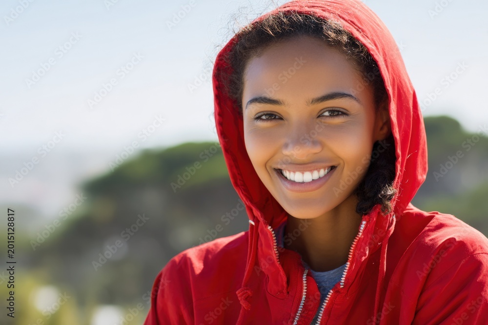 Fototapeta premium Smiling female african young closeup red hooded jacket portrait with bright teeth and blurred coastal background