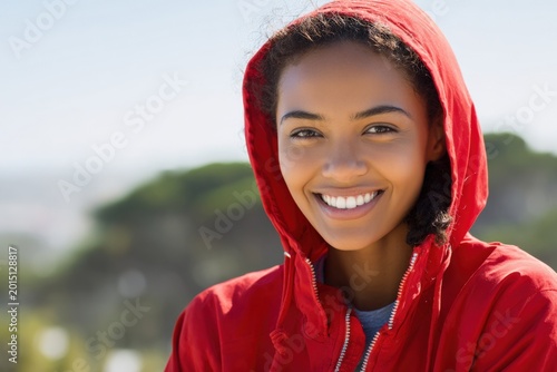 Smiling female african young closeup red hooded jacket portrait with bright teeth and blurred coastal background