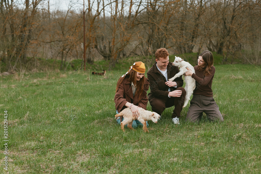 Fototapeta premium Joyful family outdoors in a park shares smiles while playing with dogs and a young goat kid, capturing playful interaction, nature, and relaxed lifestyle. Natural light highlights warm mood, casual