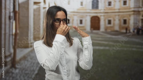 Woman pinches nose with hand and raises other palm near face in historic building courtyard; disgust avoidance.