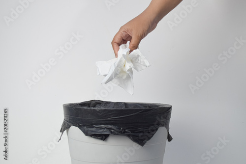 Hand throwing crumpled white tissue paper into a trash bin with a black plastic liner against a white background.