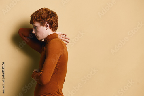 Schilderij op canvas Redhead man with curly hair wearing brown turtleneck sweater posing sideways on beige background, thoughtful and relaxed expression with hand behind head in studio photo