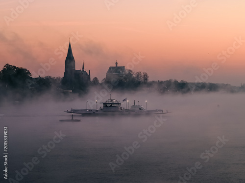 Aerial view of a ferry boat crossing the misty Meuse river with the historic church spire of Kessel silhouetted against a soft sunrise sky Kessel, Limburg, Netherlands.