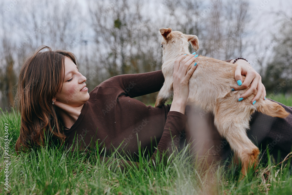 Naklejka premium Young woman with long brown hair lies on green grass holding a small baby goat in a casual sweater enjoying a peaceful outdoor moment in early spring nature.