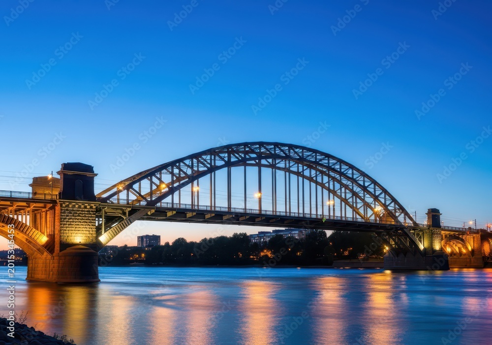 Fototapeta premium Historic stone and steel arched structures crossing a wide waterway, lit up vividly at twilight, symbolizing industrial heritage and power, massive, ironwork, riveted