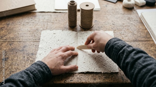 Artisan craftsman using a bone folder to crease handmade paper on a rustic wooden workbench for bookbinding
