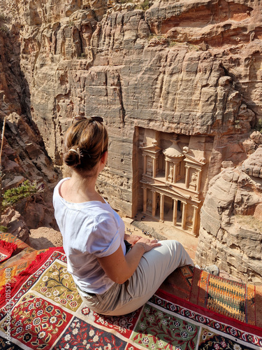 Eine junge Frau sitzt auf einem bunten Teppich und blickt auf die antike Felsenstadt Petra Khazne al-Firaun in Jordanien, eines der Sehenswürdigkeit auf jeder Reise