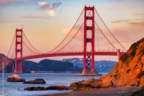 Golden Gate Bridge at sunset with warm light on towers and cliffs, calm San Francisco Bay and rocky shoreline, soft evening sky creating scenic California coastal landscape