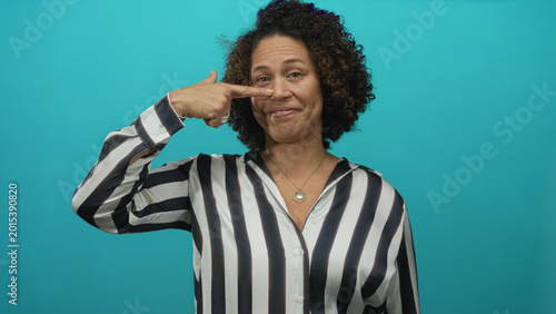 Tableau sur toile Middle aged hispanic woman wearing striped shirt pointing finger to nose in studio with teal background; confidence