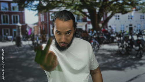 Bild auf Leinwand Man shows middle finger in street near parked motorcycles and tree by brick buildings, wearing white shirt and beard; defiance