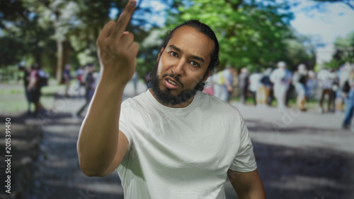 Fotografía Man with beard and white t shirt showing both middle fingers toward camera in park amid a blurred crowd; anger defiance protest