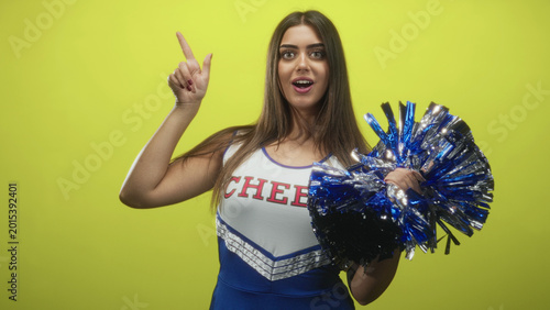 Slika na platnu Woman points finger up, mouth open, holding metallic blue pompom, wearing cheer uniform with cheer lettering in studio; team spirit enthusiasm