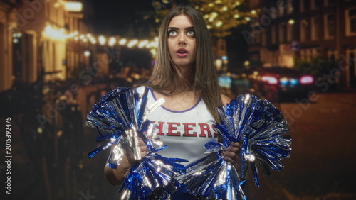 Εκτύπωση καμβά Young woman cheerleader holding blue metallic pompoms, hands visible, pouting on a street lined with string lights; boredom solitude