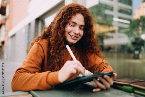 Close up of cheerful multiracial model with long curly ginger hair using stylus for digital sketching on tablet. Creative professional working on web design project, mobile application interface.
