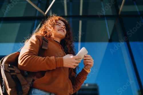 Fotografía Low angle of happy red-haired professional using smartphone against glass office building