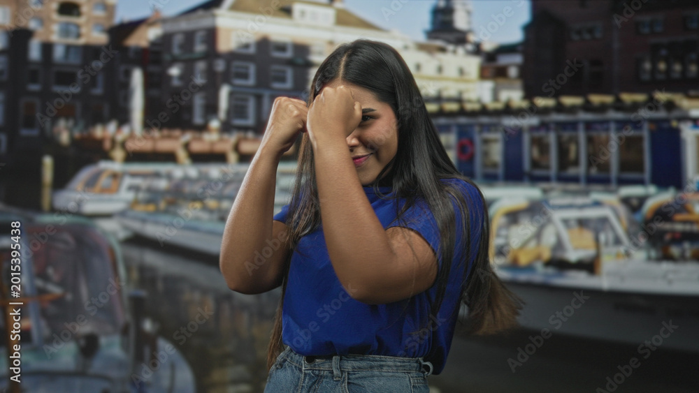 Fototapeta premium Young woman with fists raised in a boxing pose wearing a blue shirt and denim jeans in front of a city building; playful confidence.