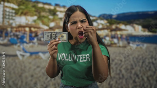 Fotografie Volunteer woman holds a hundred dollar bill and points finger while clenching fist on a beach; anger protest