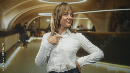 Foto Woman, middle aged blonde, hand on chest in white shirt smiling and glancing upward at airport checkin counter with arched ceiling; confidence optimism