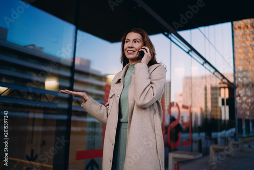 Middle aged business woman using mobile phone for international call, standing outdoors, urban glass architecture, evening light, wireless communication, technology, holding gadget, talking.