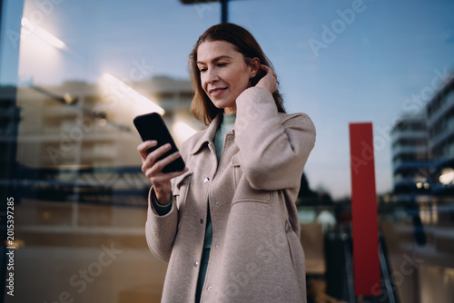 Middle aged woman browsing smartphone, reading notification or text message, standing near office building, evening urban lighting, mobile application usage, wireless data, digital nomad concept.