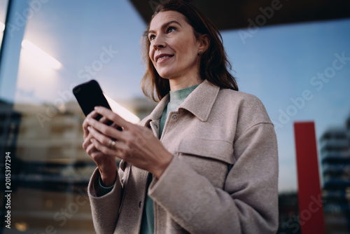 Confident lady looking away while holding smartphone, waiting for message or call, standing in urban environment, evening light, wireless connectivity, digital lifestyle, career growth concept.