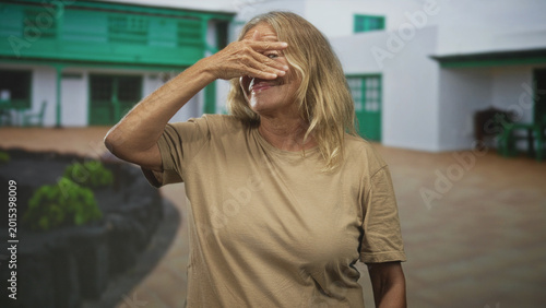 Obraz na plátně Senior woman pinches nose with hand, grimacing while standing in a building courtyard with green doors and tiled floor; disgust
