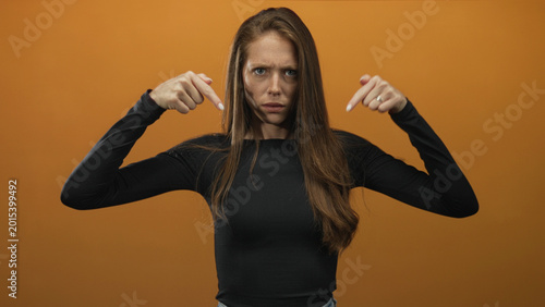 Tableau sur toile Young woman pointing both fingers down at herself in orange studio with frown and furrowed brow; accusation insistence doubt