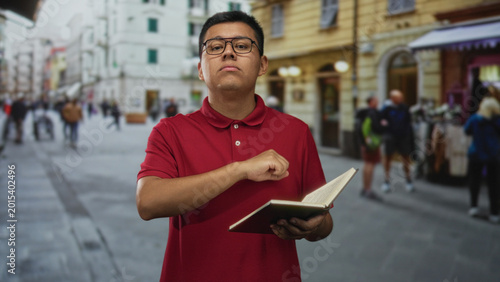 Fotografi Man in red polo shirt wearing glasses holds open book and points finger to book on street among city shops; confidence teaching