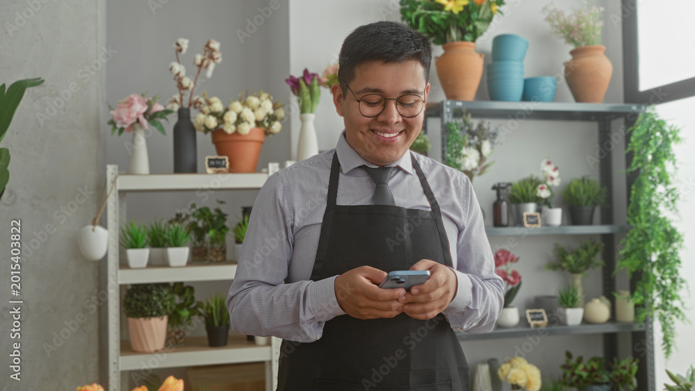Fototapeta premium Man florist in apron holds smartphone and smiles while texting in a building florist shop surrounded by potted flowers and vases; small business content.