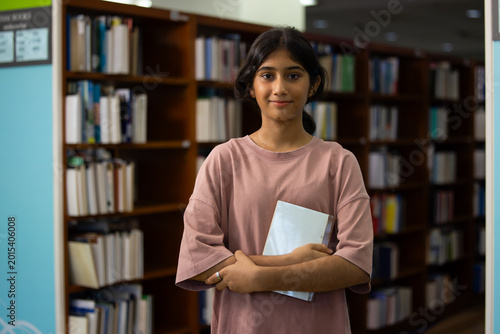 Portrait Young Indian teenage girl smiles and selects blue book. Her fingers gently touch the cover's edge. Behind her, wooden shelves hold many books. Education, Back to school