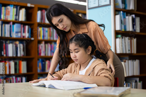 Bilde på lerret Woman guides young Asian girl through book in quiet library