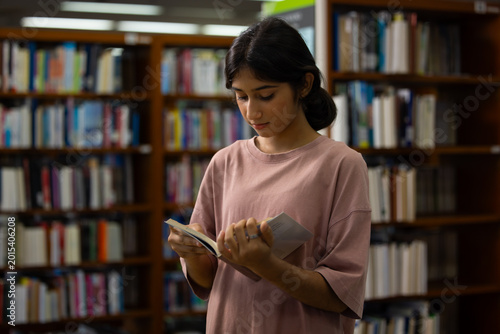 Young Indian teenage girl smiles and selects blue book. Her fingers gently touch the cover's edge. Behind her, wooden shelves hold many books. Education, Back to school