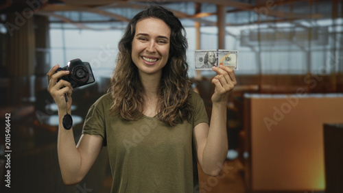 Cuadro en lienzo Hispanic woman smiling holds digital camera and crisp banknote inside brightly lit hotel building; success joy